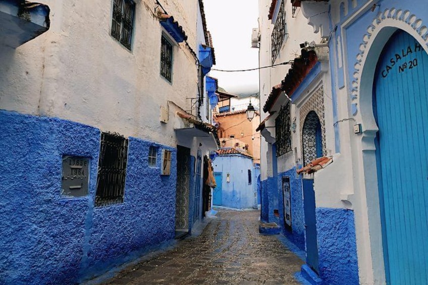 A narrow street with buildings painted in blue and white, a cobblestone path, and a blue door with the inscription 'CASA ALAH NO 20'. 4