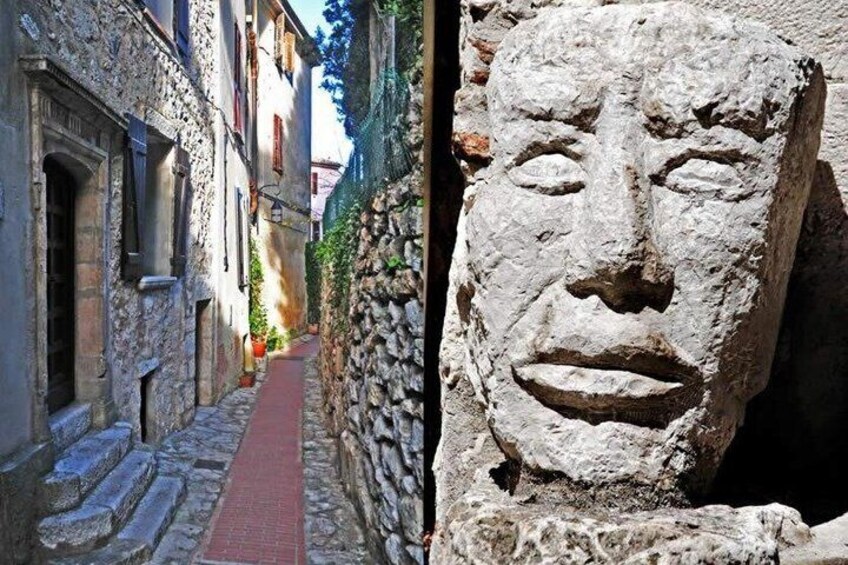 A narrow stone-paved street flanked by buildings with windows and doors, and a stone face sculpture. 2