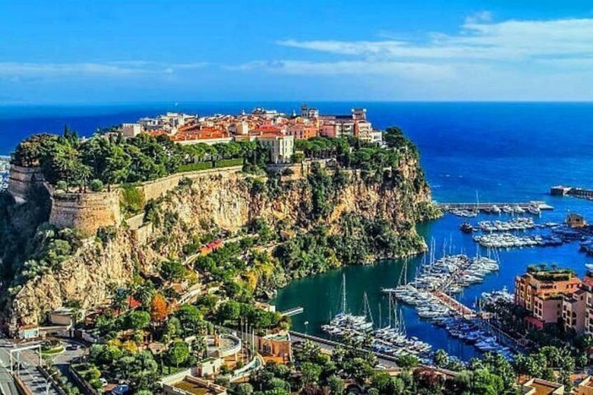Un pueblo costero con un castillo histórico en un acantilado, un puerto deportivo con barcos y un cielo azul despejado. 1