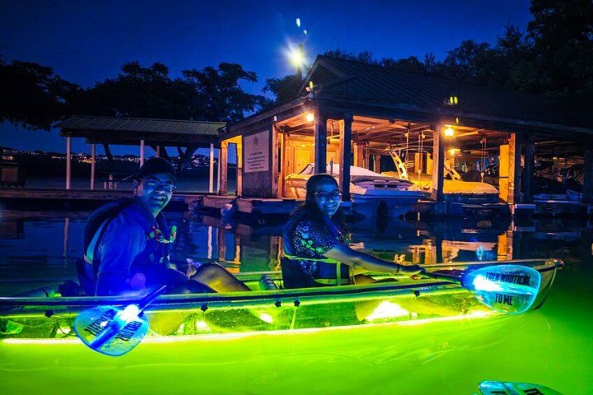 Two people in a brightly lit boat with neon lights, docked near a building with a covered area. 4