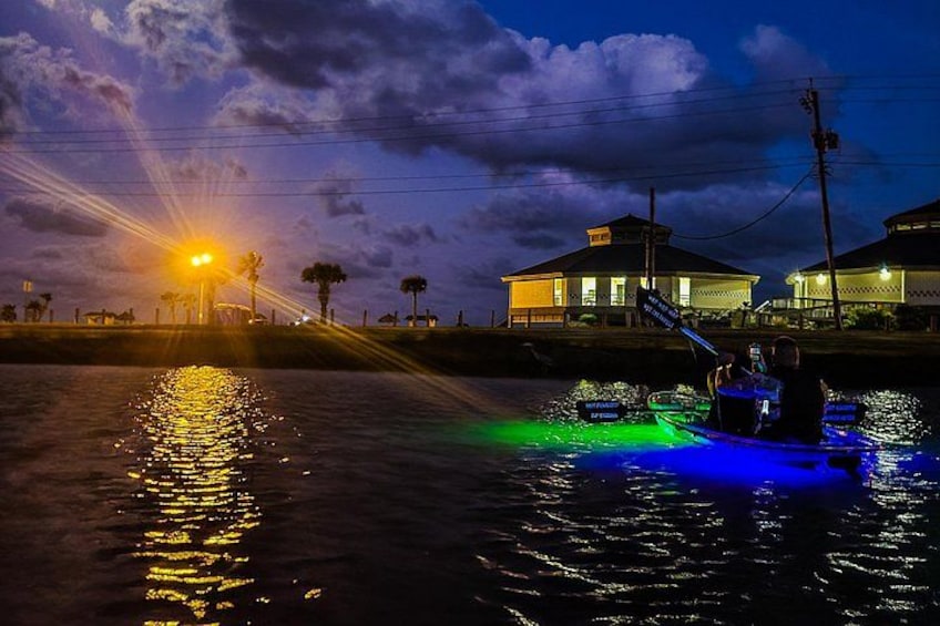 Un bote con luces azules sobre el agua, un atardecer y casas al fondo. 2