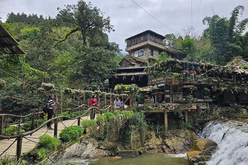 Un puente de madera sobre una cascada, abundante vegetación y un edificio tradicional al fondo. 1