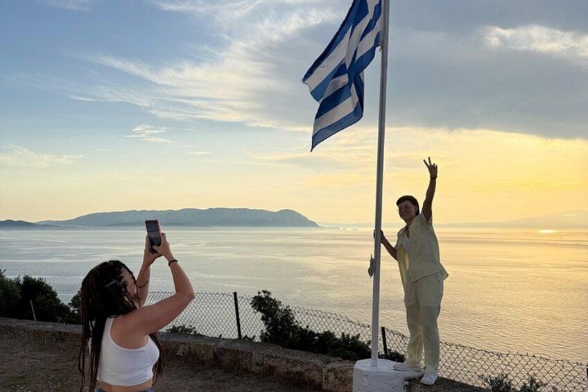 A person taking a photo with a smartphone, another person holding a flag, and a scenic view of the sea and mountains in the background. 2
