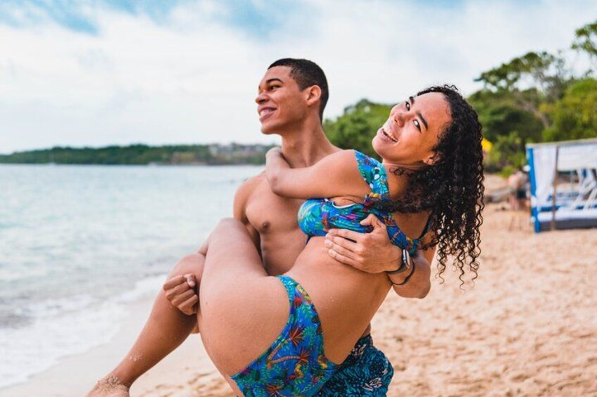 A person carrying another on their shoulders on a beach with water and trees in the background. 4