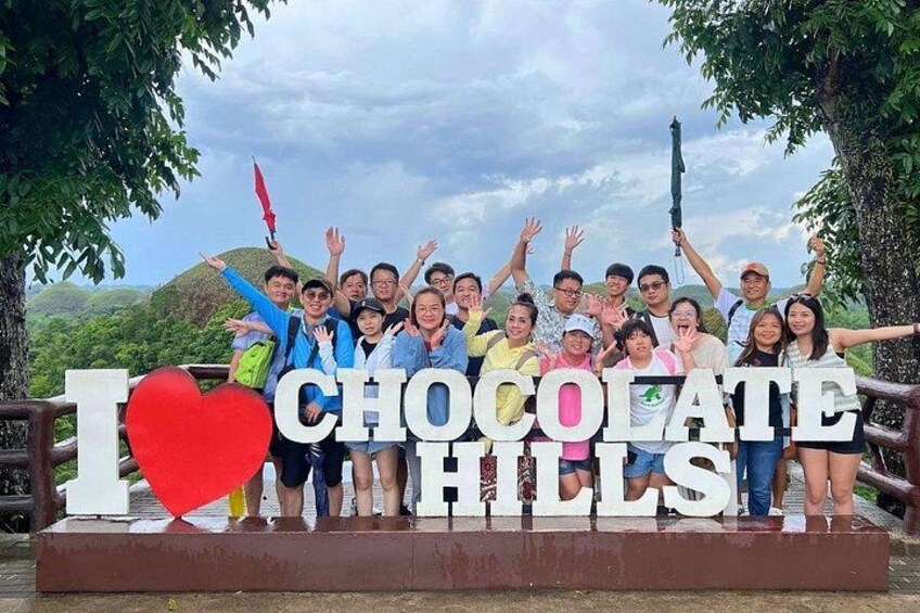 A cheerful group of tourists poses behind a large “I ❤️ Chocolate Hills” sign, with the iconic Bohol hills in the background. 1