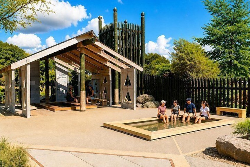 A wooden pavilion with people sitting on benches, a pool, and a fence in the background. 2