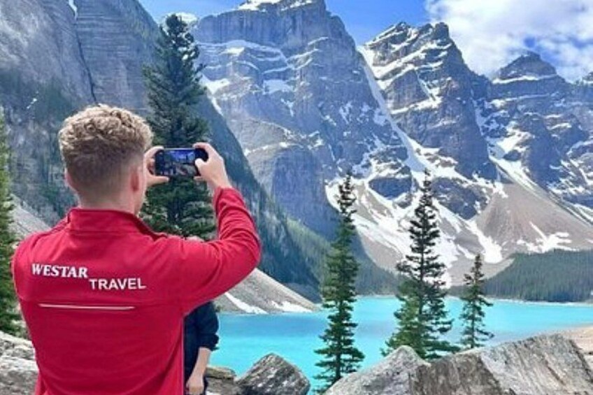 A person in a red jacket with 'WESTAR TRAVEL' on the back, taking a photo of a mountain landscape with a lake and snow-capped peaks. 4