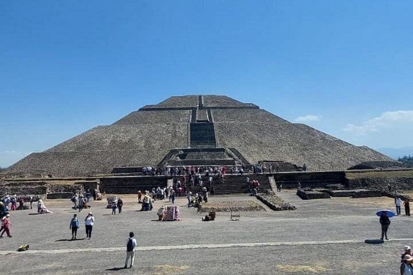 A large pyramid with a wide base and a stepped structure leading up to the top, surrounded by a large open area with people walking and standing. 3