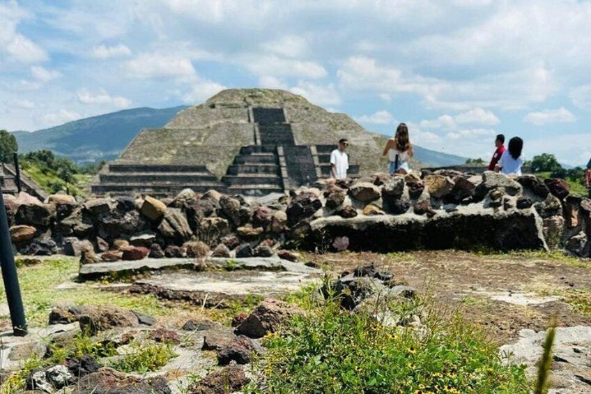 Ancient ruins with a pyramid and stone wall, surrounded by greenery and mountains in the background. 4