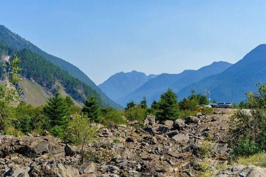 Paisaje montañoso rocoso con un vehículo en un camino de tierra, rodeado de árboles perennes y montañas a lo lejos. 4