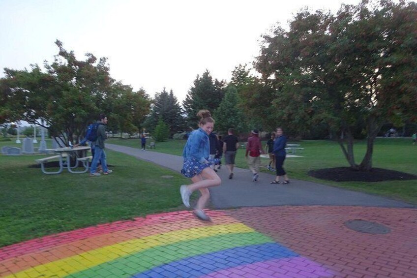 A person jumping on a rainbow-colored pavement in a park with trees and people in the background. 4