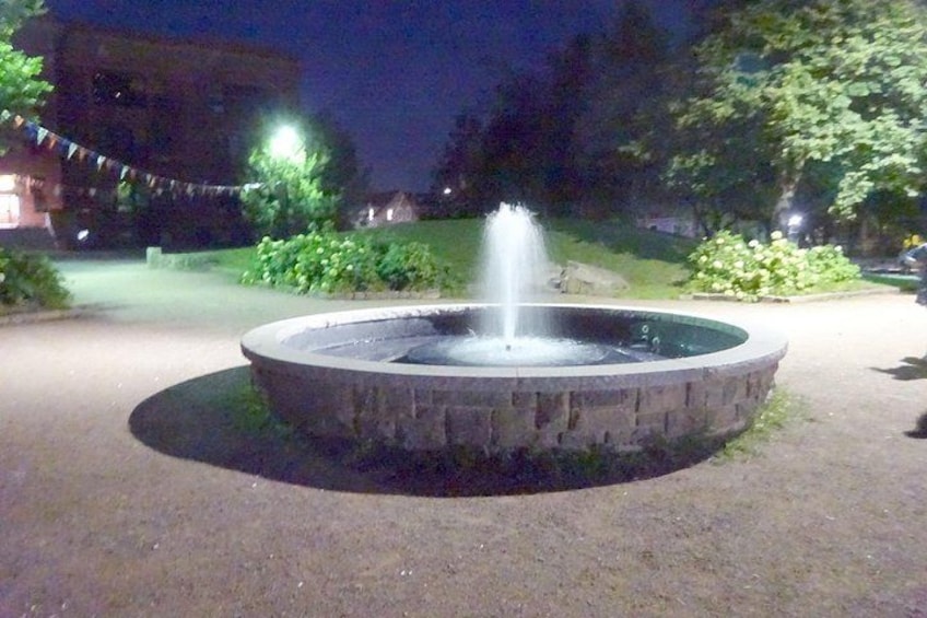 A circular fountain with water jets in the center, surrounded by a paved area with greenery and a building in the background. 3
