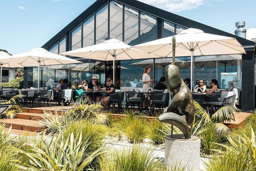 An outdoor cafe with tables and umbrellas, surrounded by greenery and a sculpture. 2