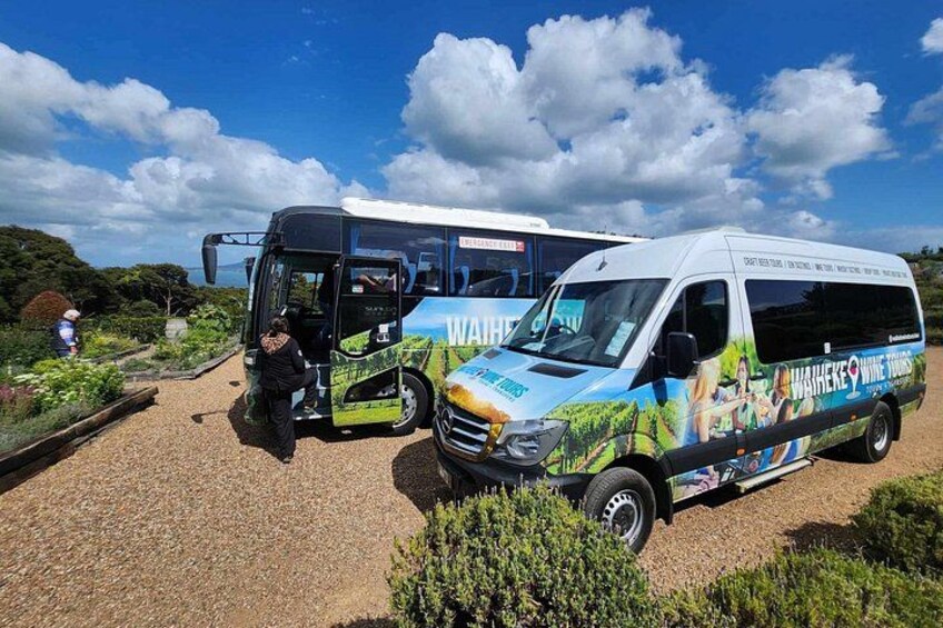 Two buses parked on a gravel surface with greenery and a clear sky in the background. 3
