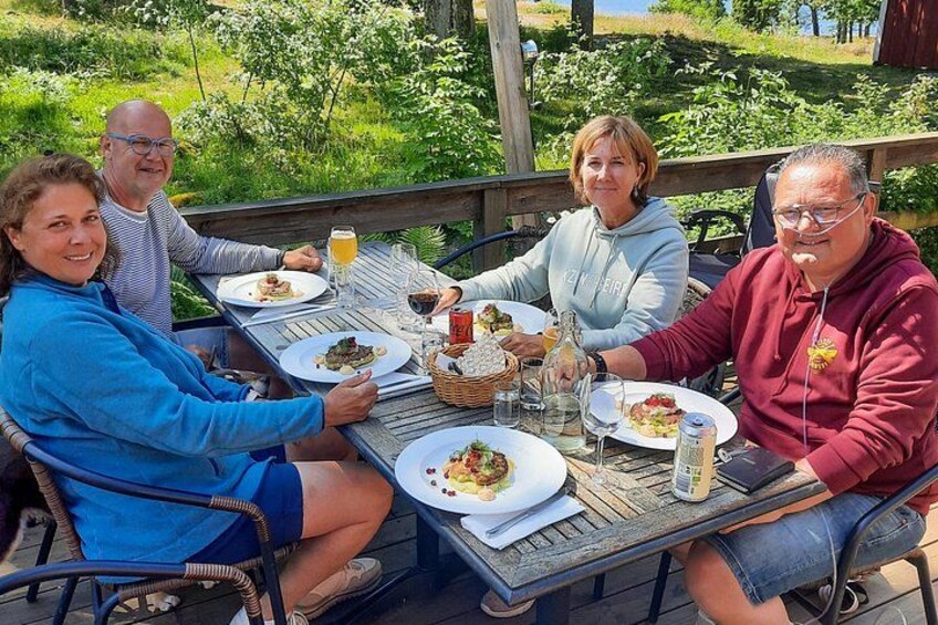 Eine Gruppe von Menschen speist im Freien an einem Holztisch mit Tellern voller Essen, Getränken und einem Korb. 2