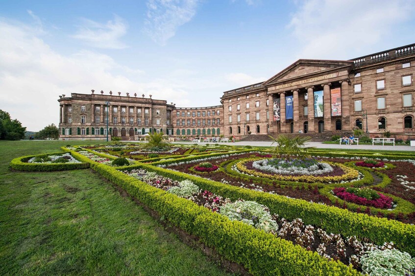 A large historic building with classical architecture and a well-maintained garden with geometric flower beds. 1