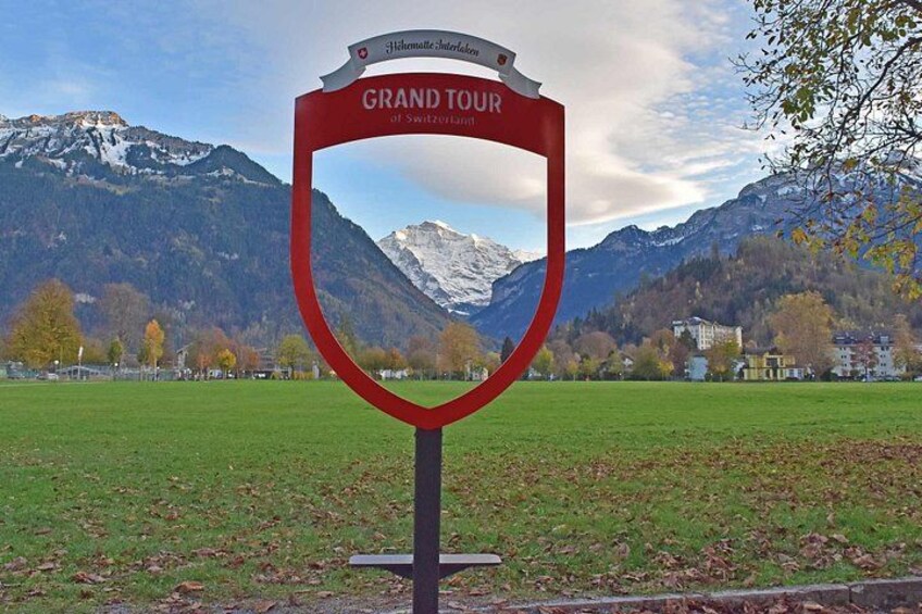 Señal roja con el texto 'Grand Tour of Switzerland', un campo cubierto de hierba y montañas al fondo. 3