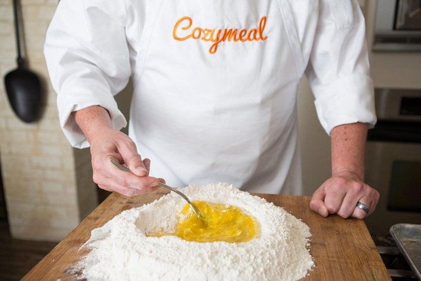 Une personne portant une veste de chef blanche, sur laquelle on peut lire le mot « Cozymmeal », est en train de mélanger des ingrédients sur une surface en bois. 1
