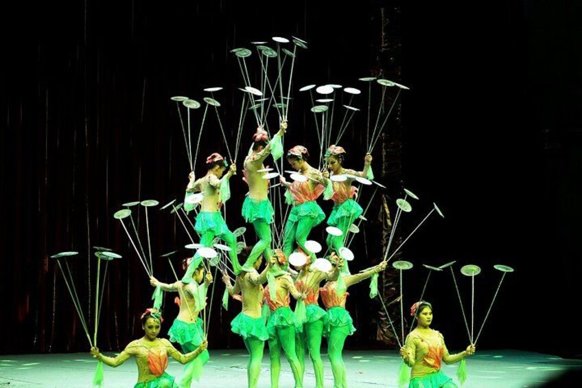 A group of performers in green costumes with props, arranged in a pyramid formation on a stage. 2