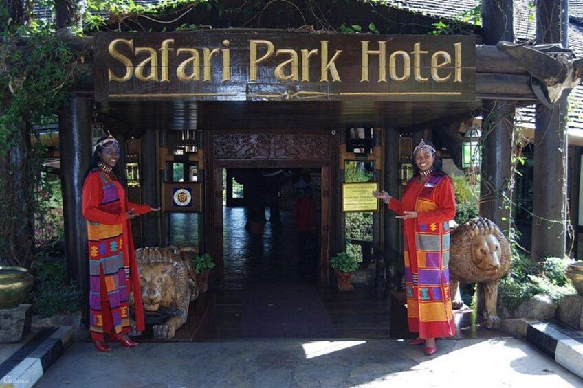 The entrance to Safari Park Hotel with a wooden sign, a lion statue, and two people in traditional attire. 1