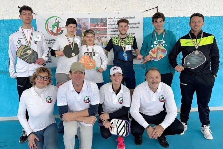A group of people posing for a photo with medals, holding tennis rackets, and wearing matching white shirts with a logo. 5