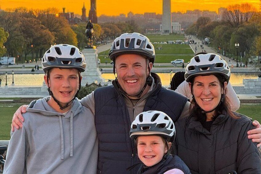 Eine Familie mit Helmen posiert vor einem Denkmal, im Hintergrund sind Wasser und Bäume zu sehen. 4