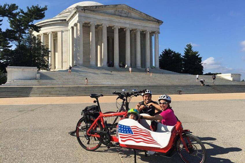 Una bicicletta rossa con una bandiera americana, una persona con una maglietta rosa e una persona con una maglietta nera e un casco, di fronte a un grande edificio neoclassico con colonne. 5