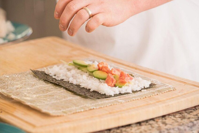 Una persona preparando sushi con arroz, aguacate y salmón sobre una tabla de madera. 2