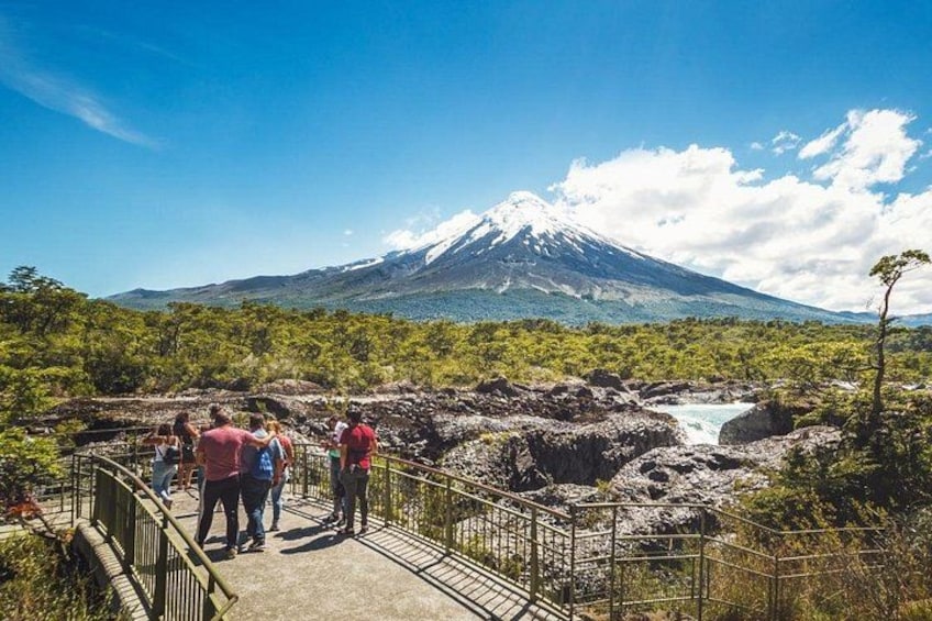 A group of people standing on a bridge overlooking a volcanic landscape with a snow-capped mountain in the background. 3