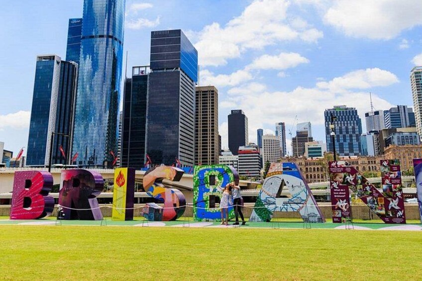 A city skyline with modern buildings, a grassy area with colorful letters spelling 'BRIBE,' and a clear blue sky. 1