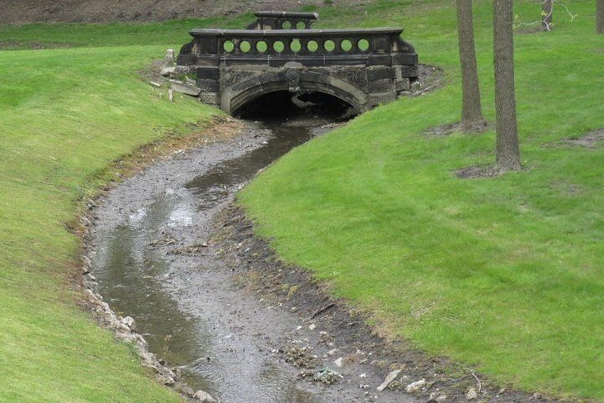 Un pont de pierre enjambe un petit ruisseau dans une zone herbeuse bordée d’arbres. 2