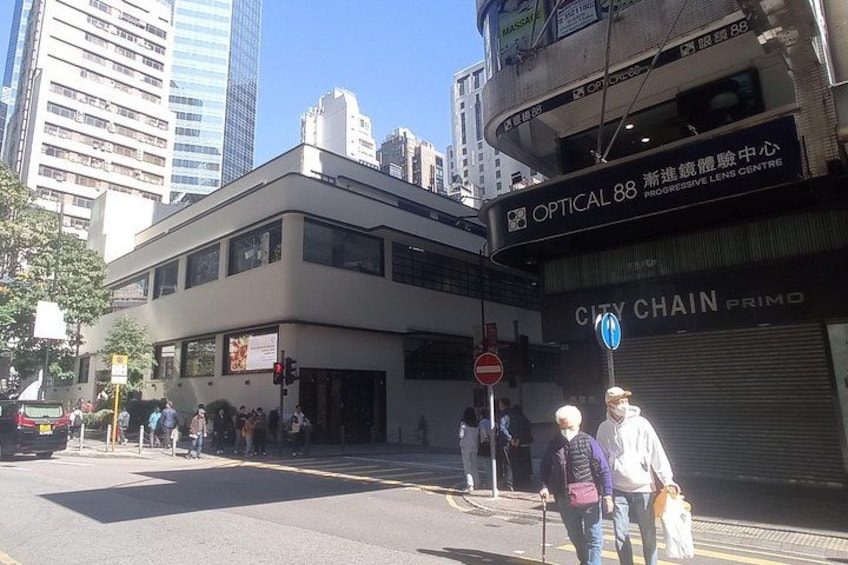 Une rue animée en milieu urbain, avec des piétons, un feu de signalisation et des immeubles commerciaux, notamment une pharmacie et un centre spécialisé dans les verres progressifs. 2