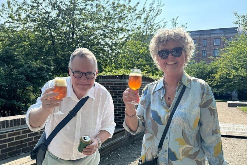 Two people holding drinks, with trees and a building in the background. 3