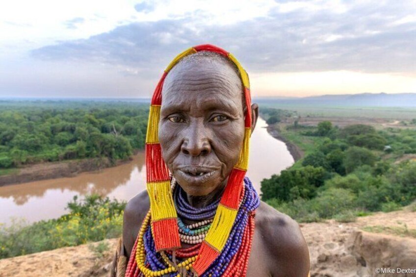 A person wearing colorful beaded necklaces, standing by a river with a lush landscape in the background. 2