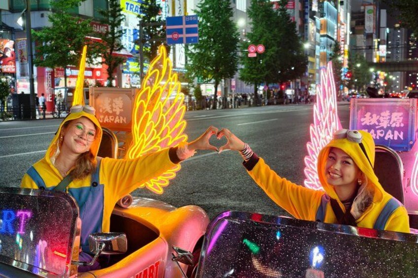 Two individuals in yellow outfits with wings, sitting in a vehicle with a neon sign in the background. 1