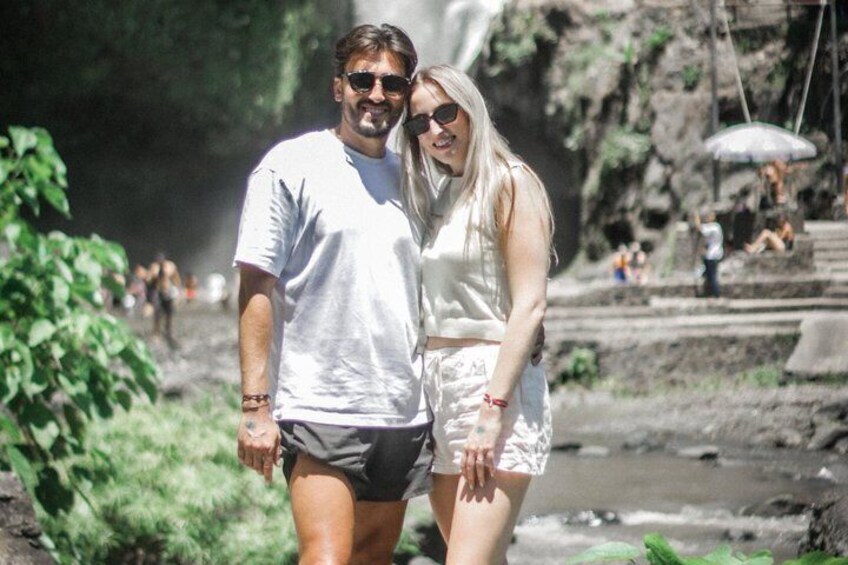 A couple walking near a waterfall with greenery and a rocky background. 2