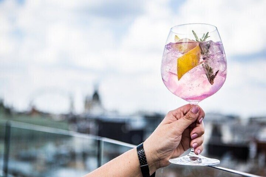 Een hand houdt een glas met een roze drankje, ijs en een plakje citroen vast, met een stadsgezicht op de achtergrond. 1