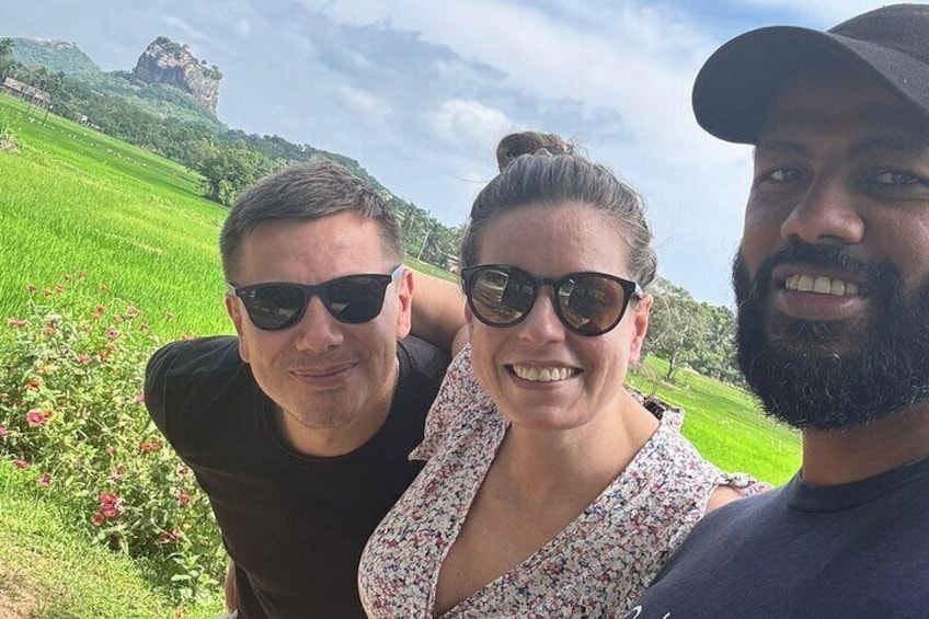 Trois personnes se tiennent à l’extérieur, devant un champ verdoyant et une formation rocheuse visible au loin. 1