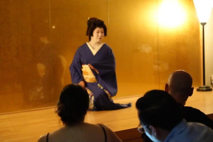 Une personne vêtue d’un costume traditionnel, assise par terre, face à un public. 2