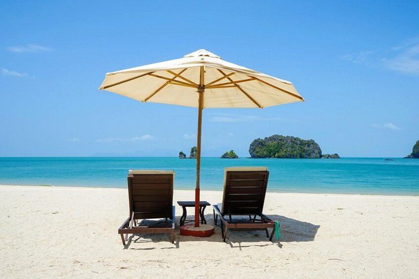 Two wooden lounge chairs and a beige umbrella on a sandy beach with clear blue water and distant islands. 3