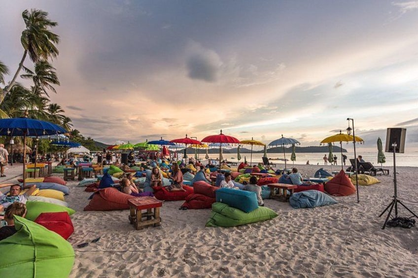 A beach with colorful bean bags and umbrellas, palm trees, and a sunset. 2