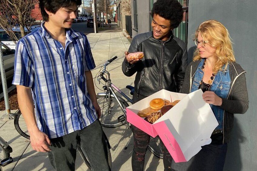Three people standing on a sidewalk, one holding a box of donuts. 5