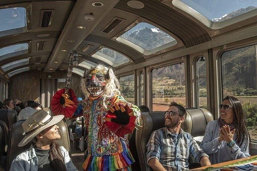 Interior de un tren con pasajeros, un artista con un traje llamativo y un paisaje montañoso visible a través de las ventanas. 5