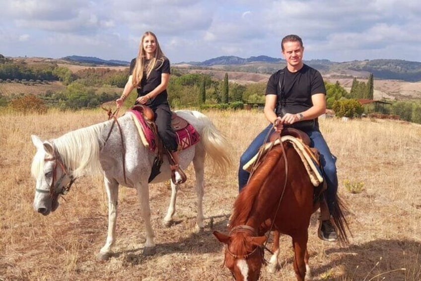 Two people riding horses in a grassy field with rolling hills in the background. 1