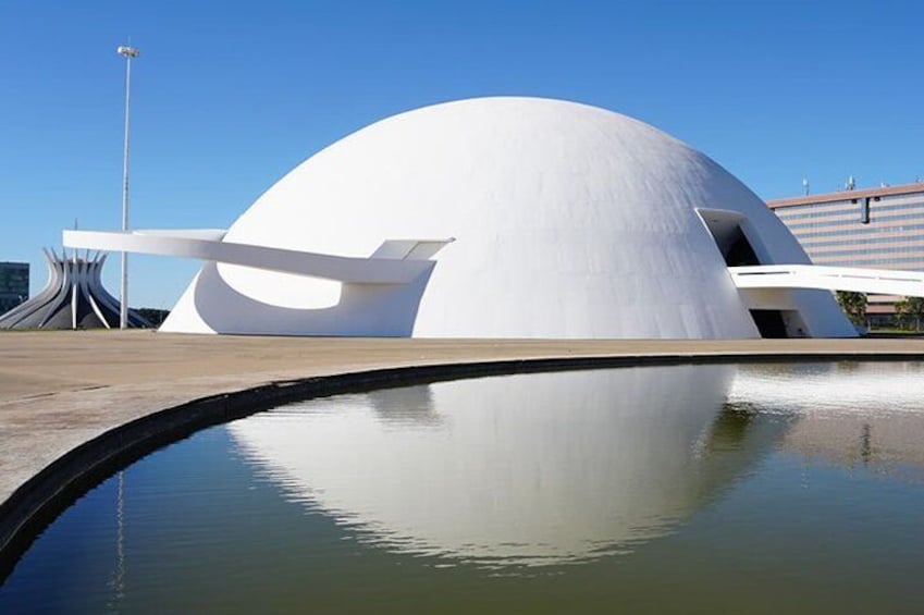 A modern white building with a unique curved roof, reflecting in a calm body of water. 4