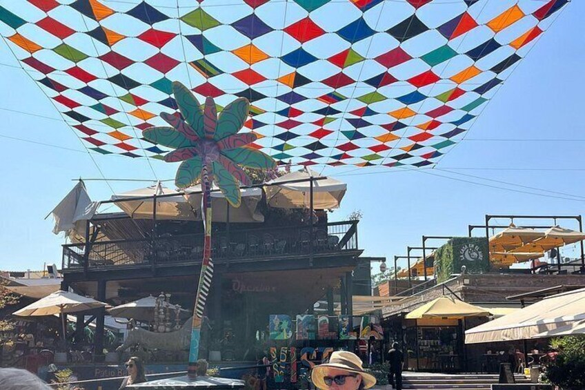 Ein farbenfroher Baldachin mit Palmenmuster, ein Gebäude mit Außenbestuhlung und ein Marktbereich. 4