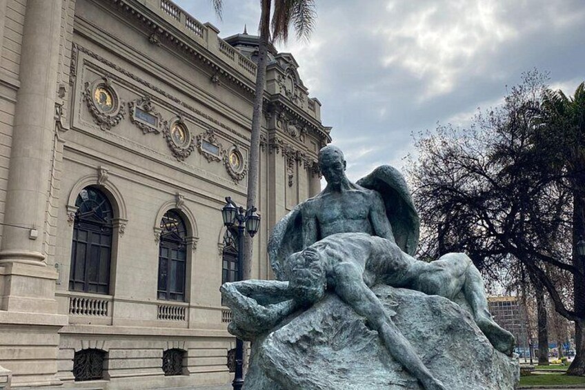 Eine große Steinstatue eines Engels mit einer menschlichen Gestalt vor einem klassizistischen Gebäude mit Rundbogenfenstern und Verzierungen. 2