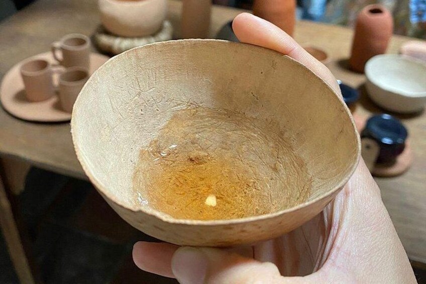 Una mano sostiene un cuenco de cerámica beige con textura rugosa y una pequeña cantidad de material amarillento en su interior, posiblemente arcilla o esmalte, en un taller con herramientas de alfarería y piezas de cerámica sin terminar al fondo. 4