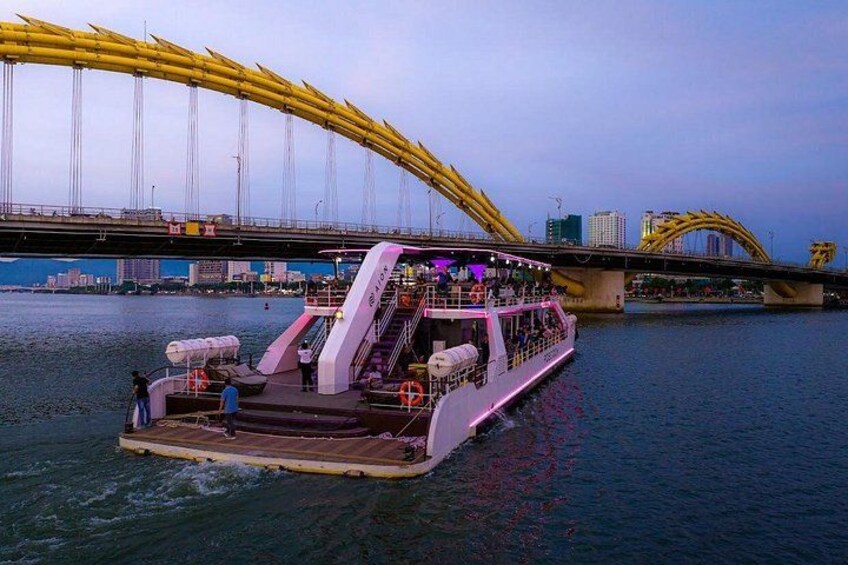 Un barco de crucero fluvial con una decoración rosa y blanca, una cubierta techada y una escalera que conduce a la cubierta superior. Al fondo, se observa un puente amarillo grande con un diseño de arco distintivo. 1