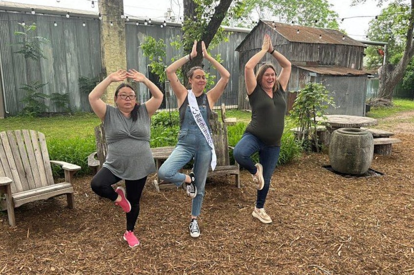 Trois personnes pratiquent une posture de yoga dans un jardin, avec une chaise en bois, une table et un bâtiment rustique en arrière-plan. 2